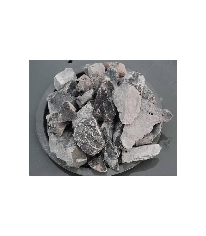 Fragmentos irregulares de carburo de calcio grisáceo, apilados en un plato metálico sobre fondo oscuro.