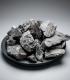 Carburo de calcio gris oscuro, trozos irregulares apilados sobre un plato negro plano, fondo gris neutro.