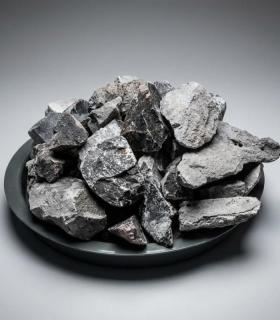 Carburo de calcio gris oscuro, trozos irregulares apilados sobre un plato negro plano, fondo gris neutro.